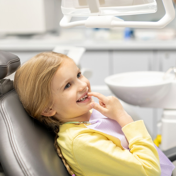 Adorable little girl showing her baby teeth to male pediatrics dentist and pointing to her smile, sitting in chair in clinic office