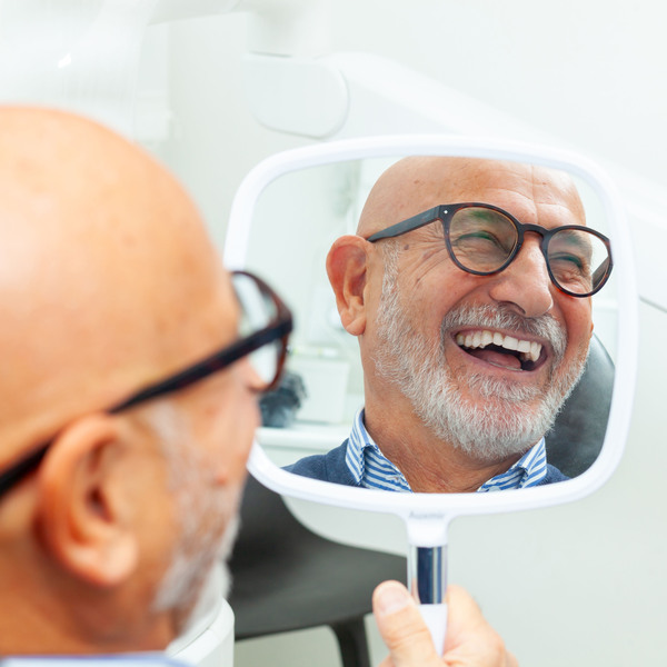 Happy senior patient admiring his perfect smile after a dental implant treatment, holding a mirror in his hand at the dental clinic