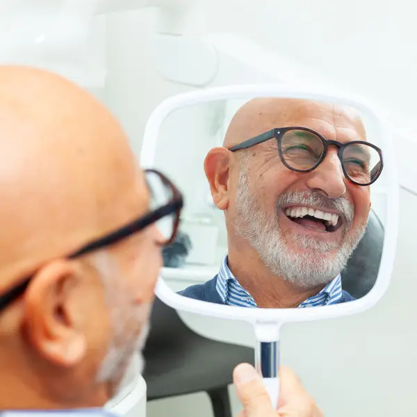Happy senior patient admiring his perfect smile after a dental implant treatment, holding a mirror in his hand at the dental clinic